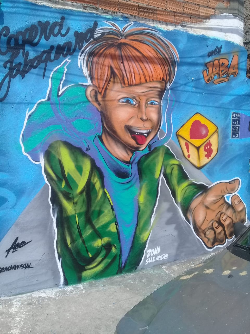 Grafite de um menino com casaco verde, cabelo vermelho , segurando um dado amarelo com o desenho de um coração. A parede está pintada de azul 